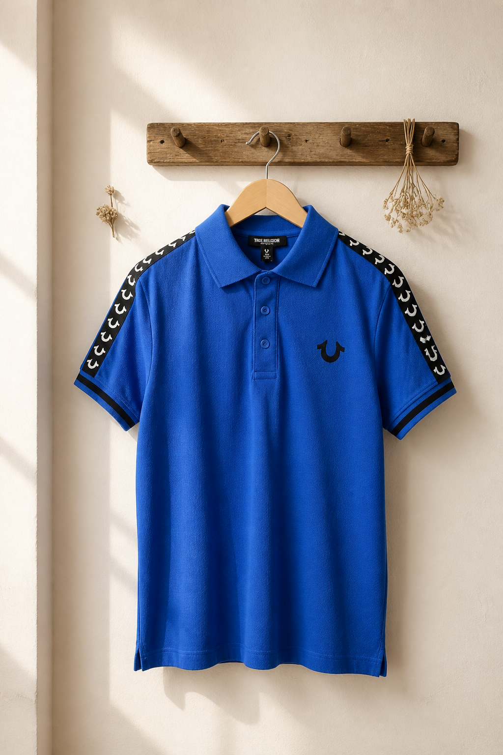 A bright blue polo shirt with black and white patterned sleeve stripes and a small embroidered logo on the chest, hanging on a wooden hanger from a rustic wooden peg on a cream-colored wall.