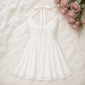A white sleeveless halter dress with a ruffled neckline and front tie details, laid flat on a white faux fur surface with lace fabric and pink roses in the corner.