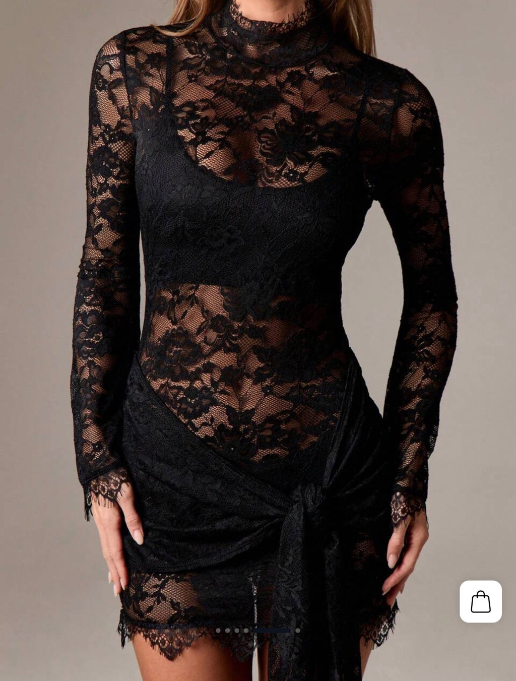 A woman wearing a black sheer lace long-sleeve mini dress with a high mock neck and a wrap-style skirt with a side tie, shown against a plain beige studio background.