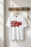 A white short-sleeve T-shirt hanging on a wooden hanger from a rustic wooden wall hook. The shirt features a large red retro-style 'Tokyo' graphic with 'JAPAN AUTHENTIC' written underneath.