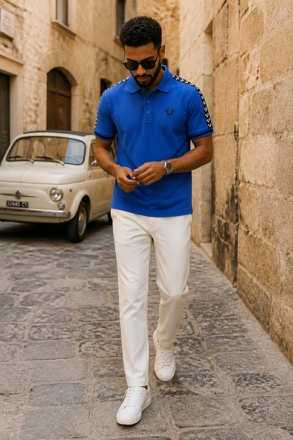 A man walking down a cobblestone street wearing a bright blue polo shirt with white shoulder stripes, white trousers, and white sneakers. A vintage cream-colored car is parked in the background against a stone wall.