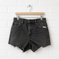 Black denim shorts on a wooden hanger against a white brick wall.
